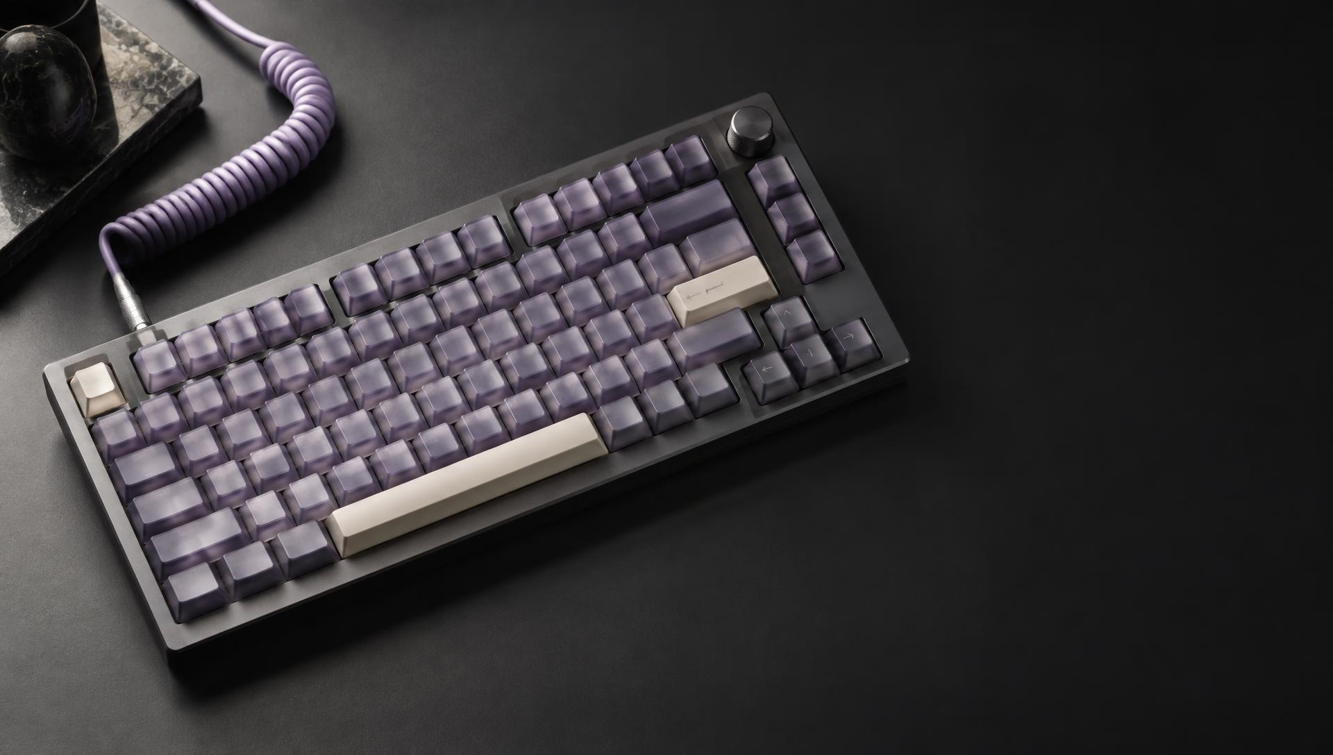 A mechanical keyboard in lilac and graphite tones on a dark desk